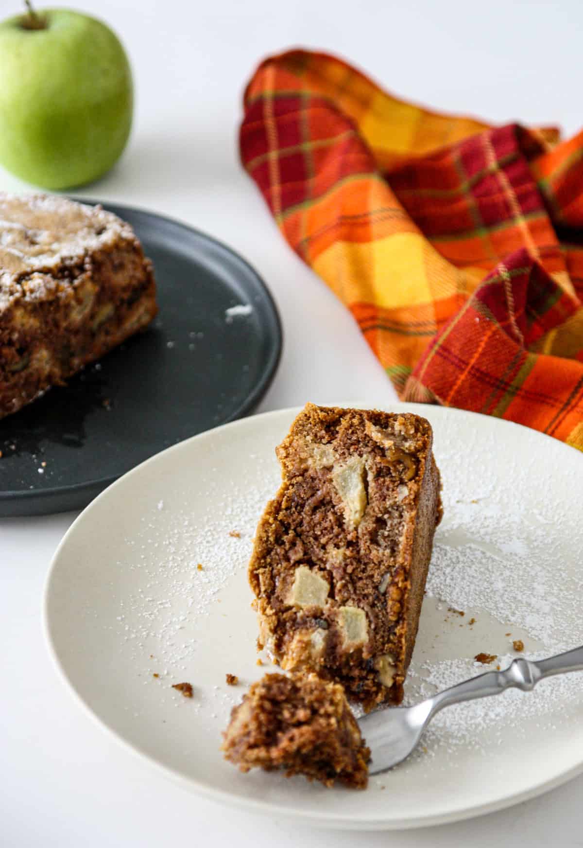 A slice a apple walnut cake on a white plate with a silver fork.