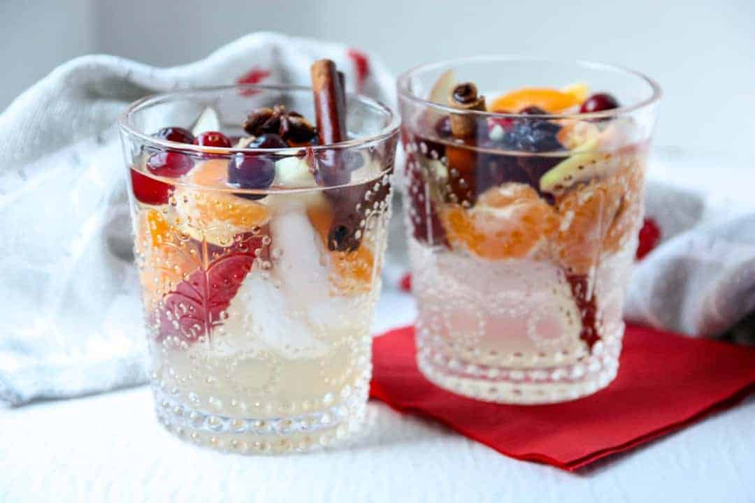 Two glasses of sangria filled with cinnamon sticks, apple slices, orange slices, and cranberries on awhite table with a red napkin.