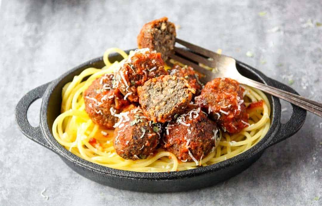 Top shot of fork holding half of meatless mushroom meatball above black bowl of spaghetti