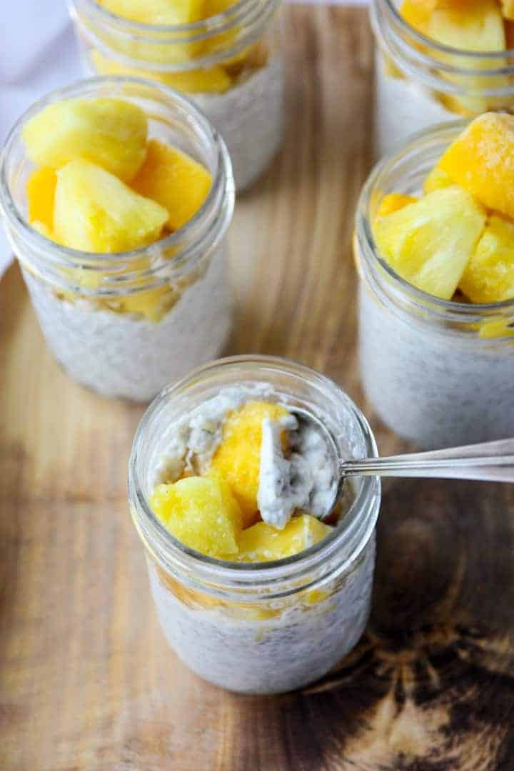 5 jars of chia pudding topped with mango & pineapple. One jar with a spoon in it.