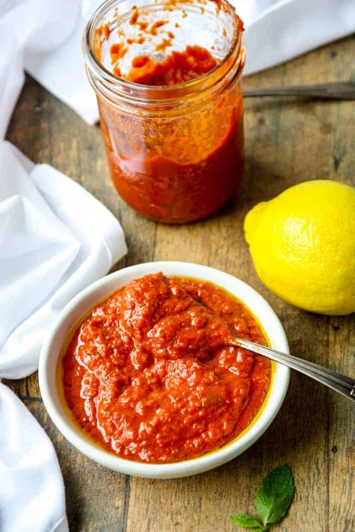 a half empty jar of harissa paste next to a white dish of harissa paste with a spoon and a lemon