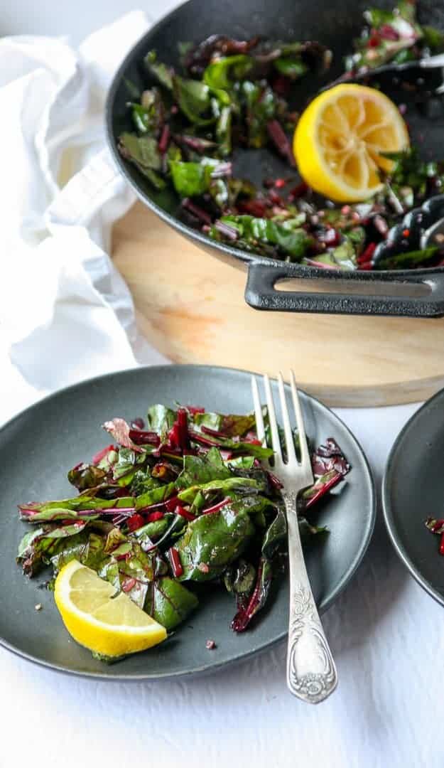 A plate of greens with a lemon wedge and a fork in front of a pan full of sauteed greens.