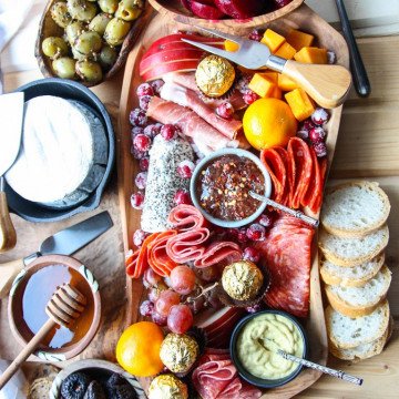 An irregularly shaped wooden charcuterie board filled with meat, cheese, olives, nuts and fruit, surrounded by crackers and bread.