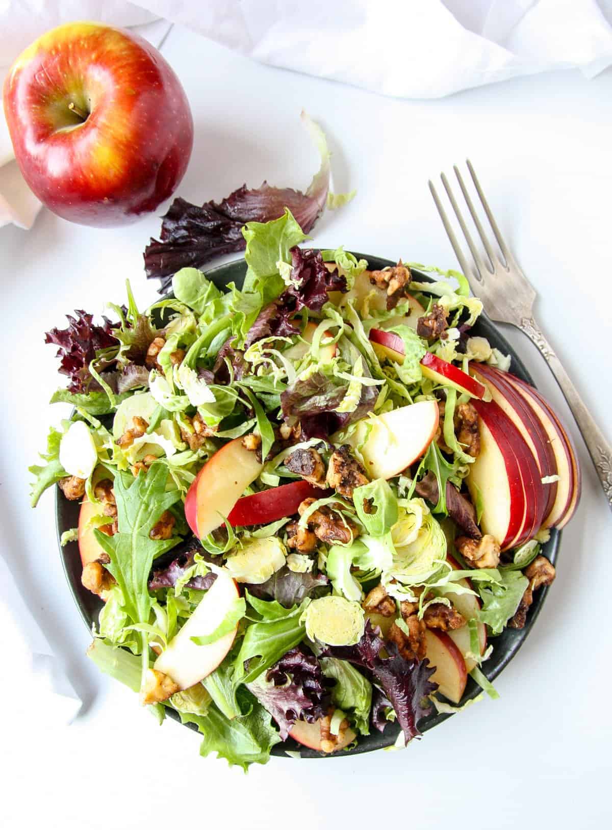 Shaved Brussels Sprouts Salad on a white plate on a white table with a silver fork, next to an apple.