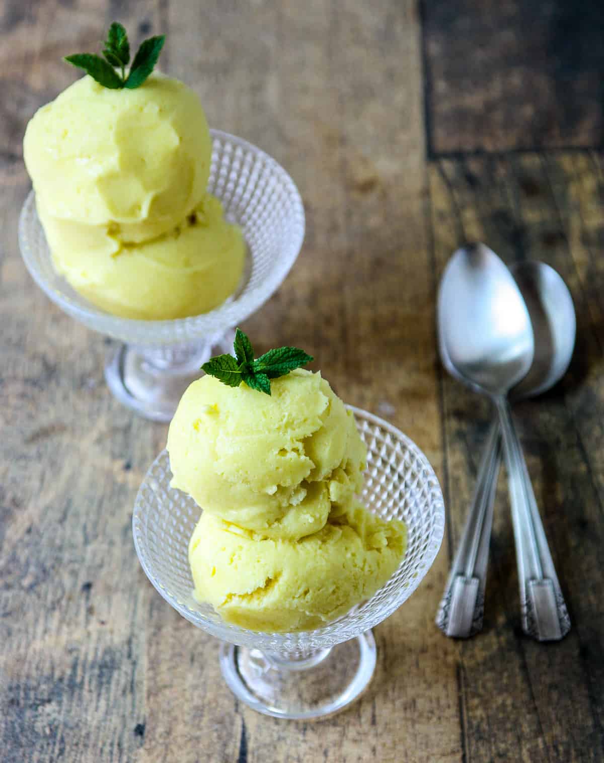 Two dishes of mango ice cream with two spoons on a wooden table.