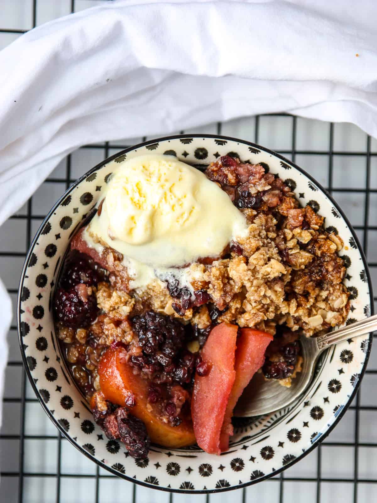 Apple and blackberry crumble dessert in a black and white bowl, topped with a scoop of vanilla ice cream.