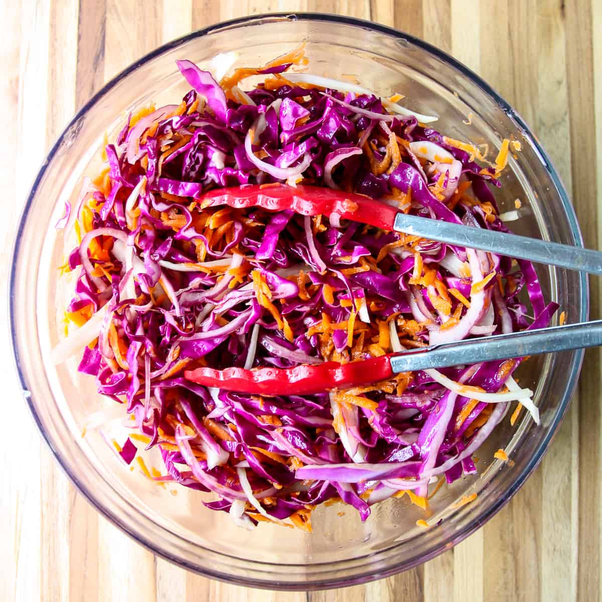 The coleslaw tossed with the dressing in a glass bowl with red tongs.