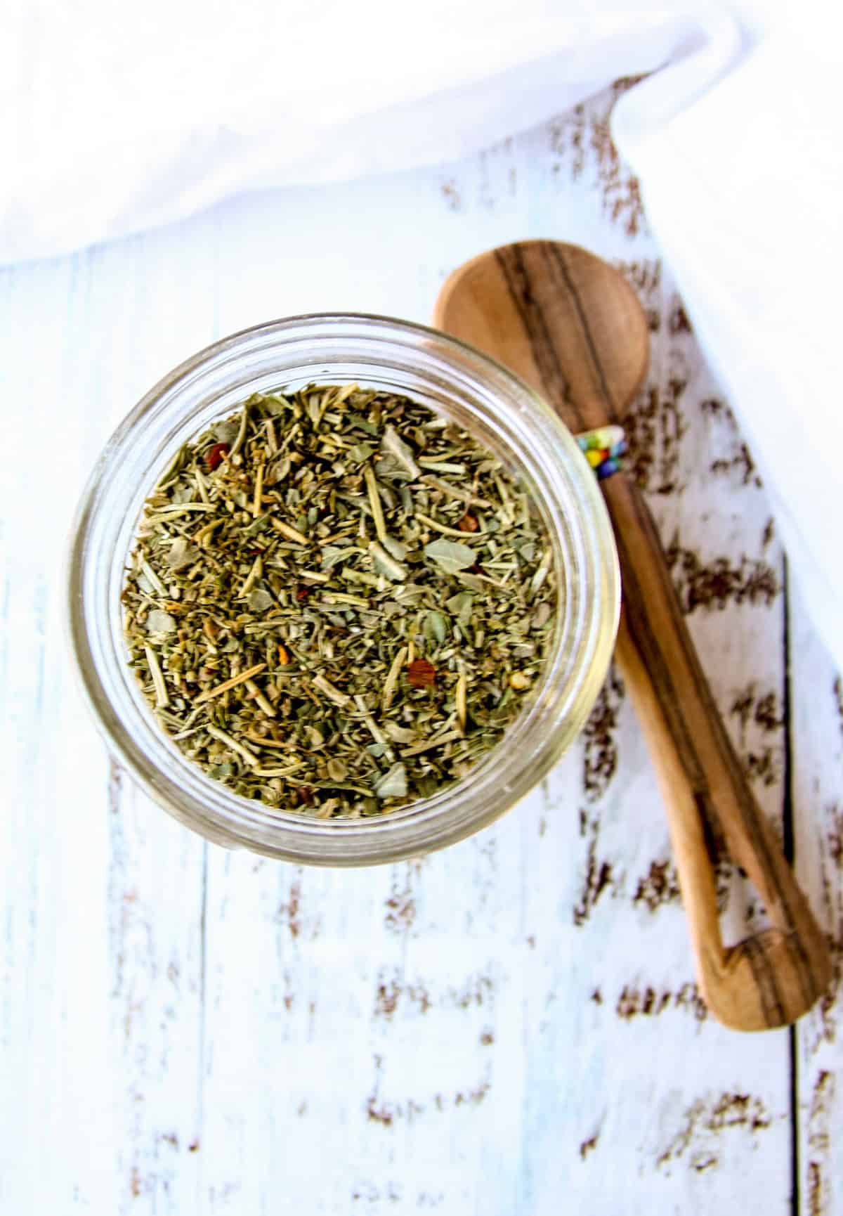 Italian seasoning blend in a glass jar next to a small wooden spoon.
