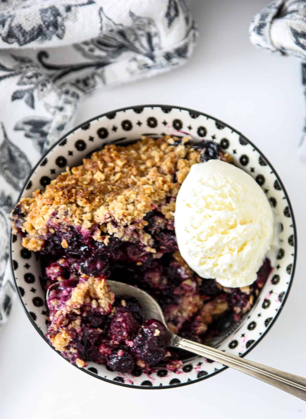 Gluten-free blueberry crisp in a black and white bowl, topped with a scoop of ice cream.
